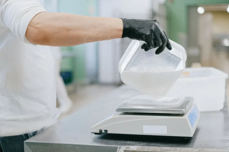 Manos con guantes pesan un ingrediente en polvo en una balanza digital: cada gramo de la fórmula cuenta en la tabla de balanceo